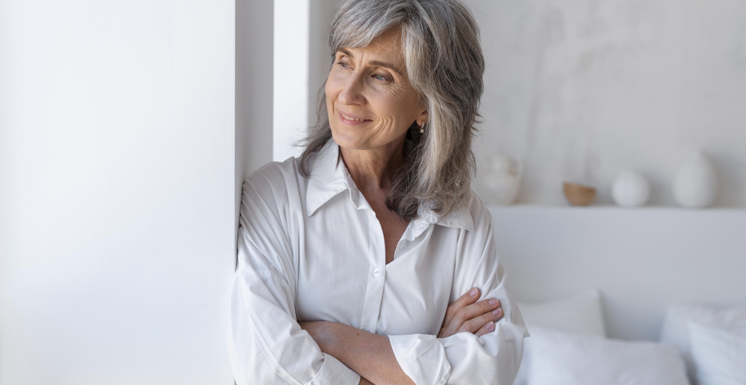 Eine ältere Frau lehnt mit verschränkten Armen an einer Wand und schaut lächelnd zur Seite.