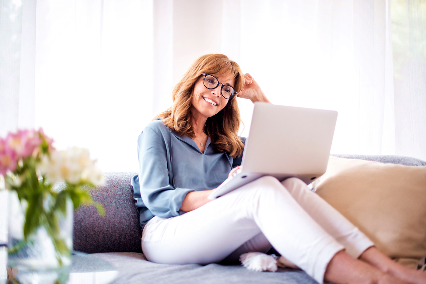 Eine Frau mittleren Alters sitzt auf der Couch und schaut lächelnd auf ein Laptop auf ihrem Schoß.